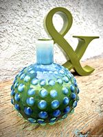 Photo showing both the olive green ampersand sculpture and the blue-green bubble effect vase together against a textured wall background on wooden surface.