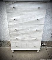 Front view of the tall off white wooden chest of drawers showing five drawers with acrylic handles and slight wear.