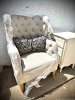 Front angled view of the neutral grey wingback upholstered chair wrapped partially in plastic. Lumbar pillow with black and cream geometric pattern is visible on seat.