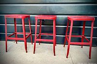 Three vintage galvanised steel bistro bar stools in red paint with visible wear and chipping on the legs and seats.