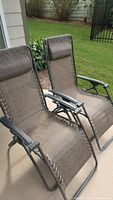 Two brown outdoor anti-gravity lounge chairs shown side by side on concrete patio. Chairs have brown fabric webbing with laced sides and black metal frames.