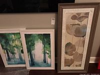 Two watercolor tree paintings in white faux wood frames with plastic coverings leaning against a wall.