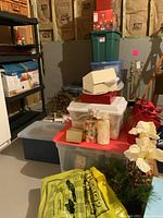 Photo showing stacked plastic storage containers holding various Christmas decorations in a basement storage area including candles, ribbons, wreaths, and other items.