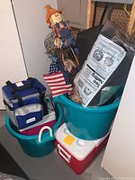 Photo showing teal plastic tubs, soft Intertek cooler, red and white Coleman cooler, scarecrow decoration, American flags and black deer netting.