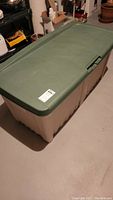 View of the large plastic storage box with green lid and beige base on the floor in basement storage area.