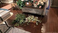 Overview of seven faux floral arrangements in baskets displayed on floor and bench, showing variety of flowers including poinsettias and leafy vines