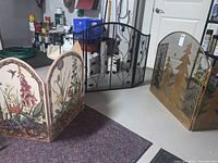 Three fireplace screens shown together on basement floor: wooden painted floral screen front left, black metal vine design back center, gold metal Christmas tree design front right.