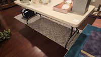 Wide angle image showing the full rectangular plastic topped folding table with metal legs placed on a patterned rug on a wooden floor. Various items on table not included.