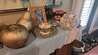 Overview of various decorative items including metallic fruits, colorful serving set, framed picture, black owl figurine, and twine sphere with shells on a table