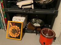 Photo showing George Foreman grill on top shelf, Oster bread maker, pressure cooker, warming tray, and framed food photo on bottom shelf in basement storage.