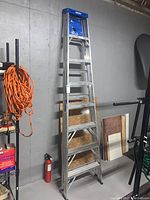 Full view of Werner 8-foot aluminum step ladder standing folded against basement wall with shelf and other tools nearby.