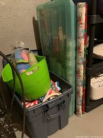 Gray storage tote with a bright green plastic beverage tub inside, and partially visible translucent green plastic case holding wrapping paper rolls stacked upright behind the tote. Some wrapping paper rolls leaning upright next to case.