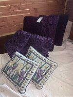Seven pillows in a group showing texture and variety: two tapestry pillows with flowers and butterflies, two purple petal pillows, three faux fur pillows (two purple, one cream).