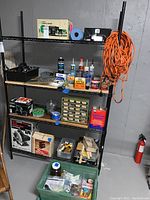 Photo showing black shelving unit with various items including boxed Craftsman circular saw, wooden pieces, several cans and tubes of adhesives and cleaners, Frog tape, joint tape, and trays. Bottom shelf has a green plastic bin with assorted small items.