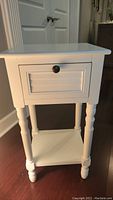 Full view of the small white side table with one drawer and lower shelf showing turned legs and tabletop.