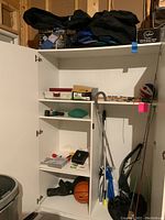 Full view of white storage cabinet containing fishing rods, reels, net, tackle box, hunting cutlery, sleeping bag, sports balls, and accessories.