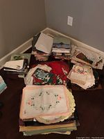 Stacks of assorted table linens including folded and packaged tablecloths, placemats, seasonal napkins, embroidered pillowcases, and doilies on the floor.