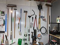 Wide view of assorted yard and garden tools hanging on pegboard including rake, pitch fork, shovel, axe, hand saws, hammers, caulking gun, wire coil, and various small tools on the right side.