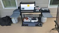 Weber Genesis Gold gas grill with cooking grate and warming rack visible, two white propane tanks under side shelves, cookbook, accessories arranged on and around grill, black grill cover on ground beside it, and a grill mat underneath.