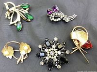 Five vintage brooches including a green rhinestone gold-tone bow, a silver-tone purple and clear rhinestone floral style, a gold-tone bow with pearls, a black and pearl stone floral brooch, and a gold-tone brooch with a red stone.