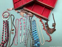 Wide view showing multiple necklaces with various bead styles and colors including red coral, white, pink, blue, and dark beads alongside a wood jewelry box with red lining and compartments.