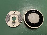 Front view of the Nest Smart Thermostat showing the circular black face with some scratches and the white mounting plate with terminal labels.
