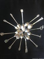Top view of all 13 sterling silver souvenir collector spoons arranged in a circle, showing varied handles and polished bowls