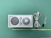 Front view of Tivoli Audio Model One radio showing speaker grille, analog tuning dial, volume and band knobs, with power cord and antenna wire. Lumbar ruler for size reference included.