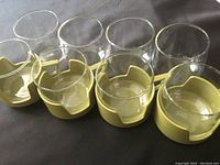 Four clear glass drink cups in yellow plastic holders with notch and handles, arranged on flat surface.