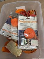 Plastic bin with multiple orange ratchet tie down straps, some in original packaging marked 'Professional Type Ratchet Tie Down', others rolled and loose.