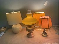 Four table lamps arranged on a surface showing three with brass bases and one with ceramic base, with varied fabric and lantern style shades.