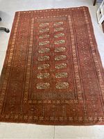 Full view of rectangular wool area rug with reddish-brown base color and repeated oval floral pattern in center, surrounded by patterned borders. Visible signs of wear.