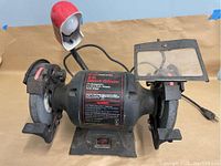 Front side view of the Craftsman bench grinder showing the work light on the left, grinding wheels, tool rests, and eye shield over right wheel.