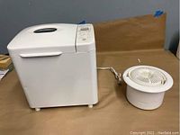 Side view of the white bread maker plugged in showing its main body and the citrus juicer placed next to it.