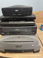 Stacked stack of electronics showing Sega Saturn gaming console on top, Sony DVD/CD player, Macintosh PowerBook G3, Toshiba VHS recorder/player, and Microtek ScanMaker 900 scanner on bottom.
