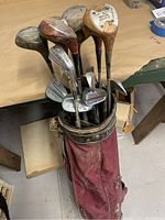 View of the full set of golf clubs in worn red golf bag, showing wooden woods and metal irons with visible wear and tear.
