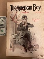 Cover of The American Boy November 1913 featuring an illustrated boy seated by a wooden bench with a pipe, holding a chicken. Shows a dollar bill for scale to indicate size.