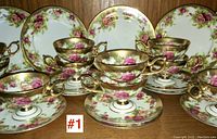 Front view showing 12 footed cups and saucers in rose floral and gold trim pattern with 10 side plates behind