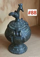 Front angle showing the rounded body, decorative patterned surface, and bird-shaped finial on the lid.