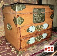 Front-side angled view showing wooden box with brass corner protectors, stone carvings on drawers, brass handles and decorative lock plate.