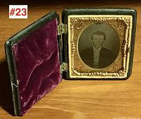 Black hinged gutta-percha photo case opened showing hinged interior lid lined with deep purple velvet and inset with a mounted Daguerreotype photo of a man in formal attire under an embossed metal mat with ornate floral designs.