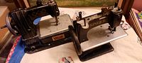 Pair of antique child's tin toy sewing machines side by side on a cloth surface, Vulcan on the left and Singer on the right, showing overall condition and scale.