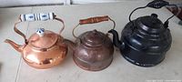 Three metal tea pots arranged side by side on a surface, showing front and side views. Copper pots are bright with aged patina on one; black pot has a ribbed body and coiled handle.