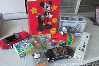 Collection of vintage toys including Disney Mickey Mouse items, diecast cars, and a boxed model kit on a white table.