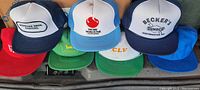 Photo showing assortment of vintage trucker hats with visible logos including Ridinger Bros. Painting, The 1982 World's Fair, Becker's Sunoco, John Deere, and CLV in mesh back snapback style.