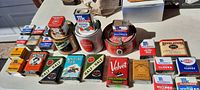 Wide shot showing all 26 vintage tins arranged on a table with sunlight highlighting colors and labels. Tins include various McCormick spice varieties, tobacco tins, and other country store style collectibles.