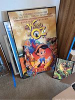 Framed posters leaning against a wall including a large Pinocchio special edition Wizard of Oz poster and a smaller dinosaur poster from The Lost World.