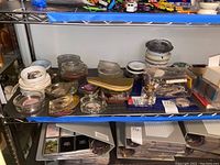 Wide shelf of assorted ashtrays in glass and ceramic with printed logos and designs alongside can openers and decorative plates.