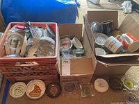 Three boxes and crates filled with assorted ashtrays and mugs from various companies and travel destinations.
