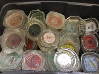 Overhead shot of plastic bin filled with multiple glass ashtrays, showing various designs and logos including Phoenix Motel and bowling lanes.